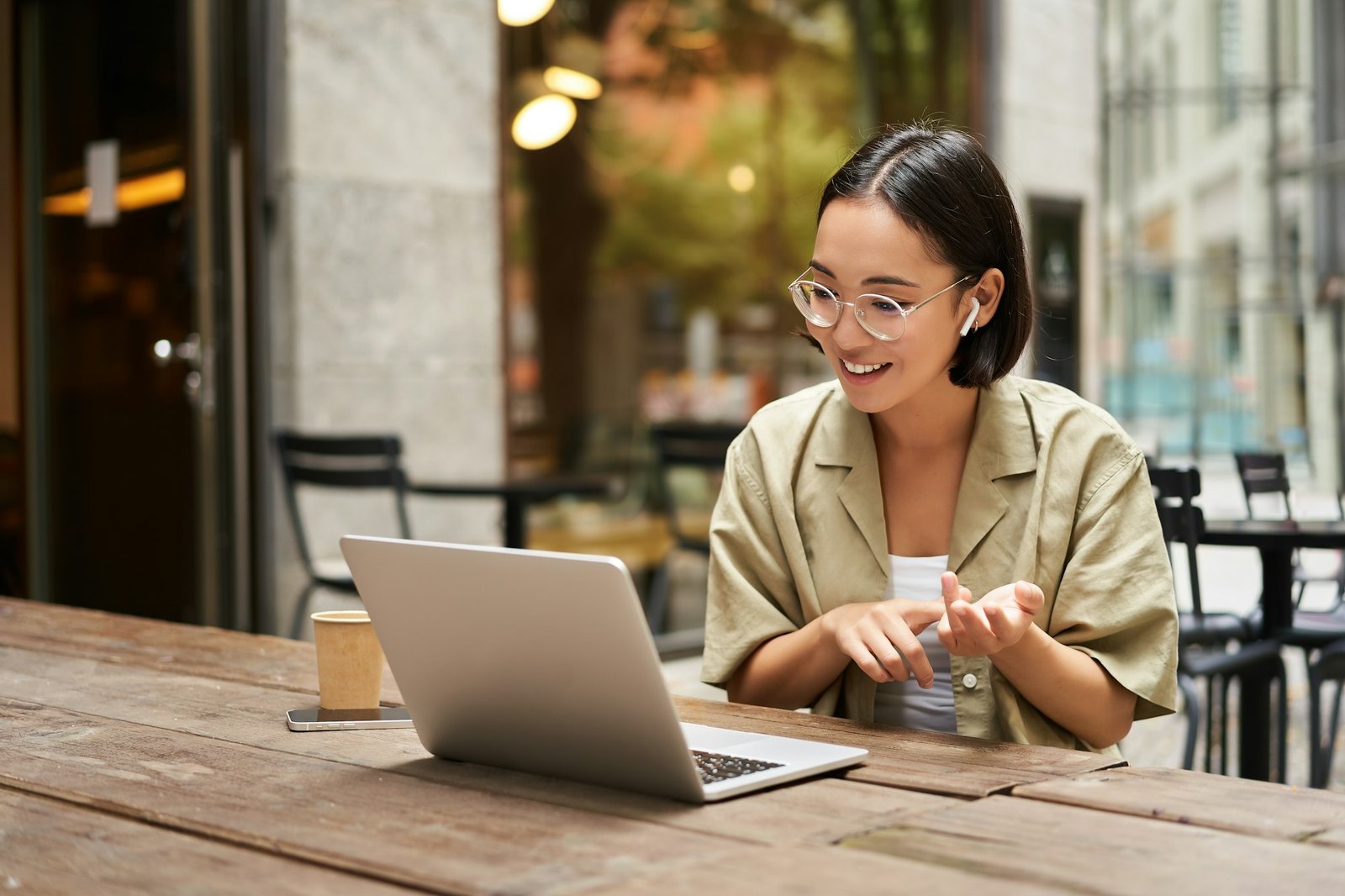 Young woman sitting on online meeting in outdoor cafe, talking to laptop camera, explaining